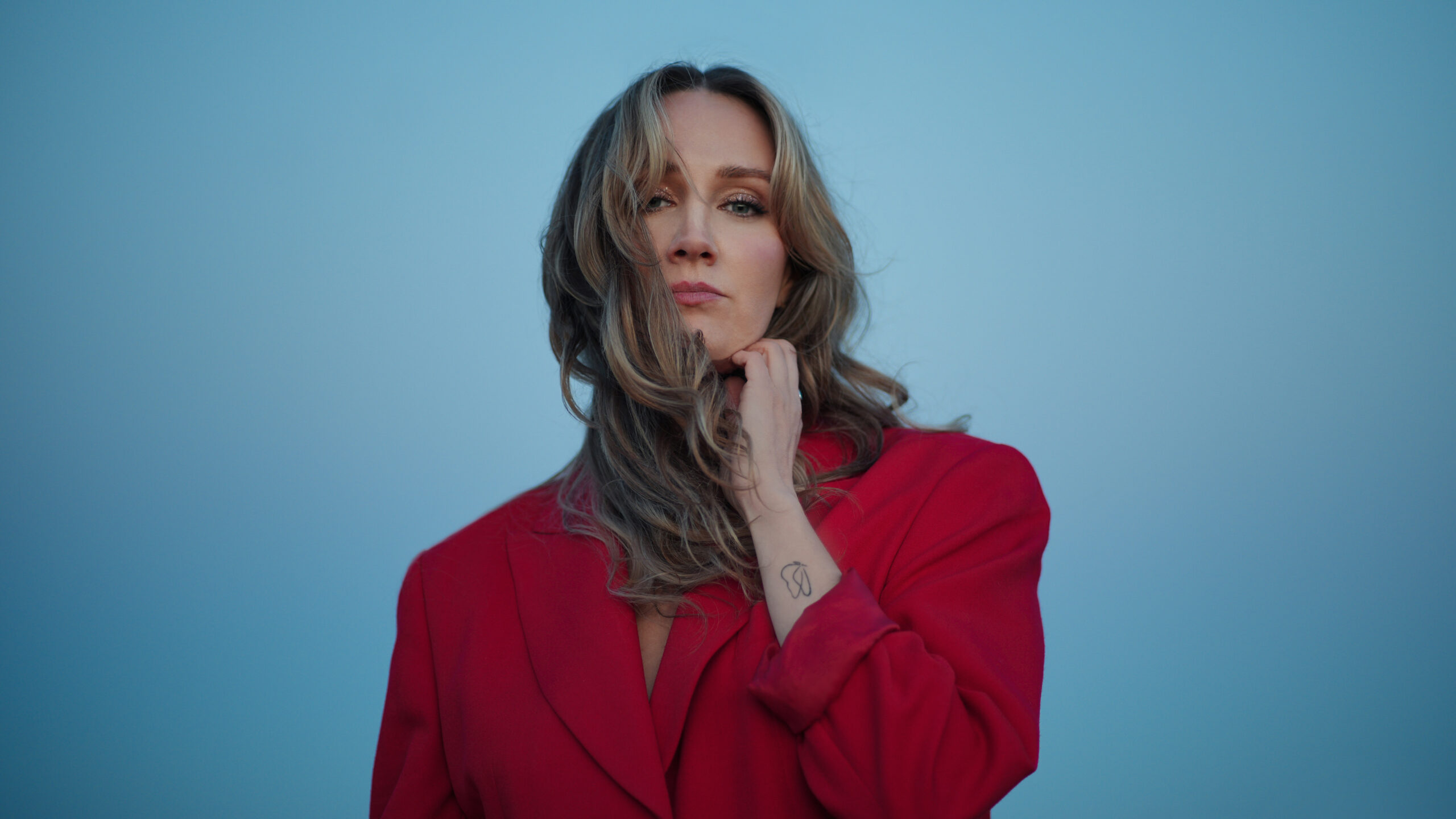Rachel Fenlon wearing a red blazer with her hand held up to her neck with a blue background